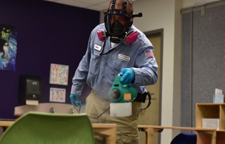 17th Civil Engineer Squadron pest controller Omar Martinez, walks around a classroom dispersing a disinfectant mist on all surfaces at Goodfellow Air Force Base, Texas, March 30. The chemical used is reportedly 99.99 percent effective at bonding to germs on the surface and killing them within the dwell time of 20 minutes. (U.S. Air Force photo by Senior Airman Seraiah Wolf)