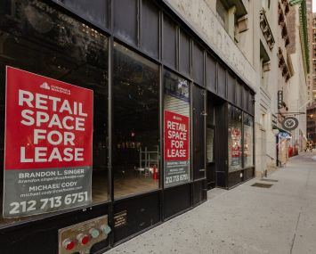 Empty storefronts with leasing signs on them along an empty sidewalk.