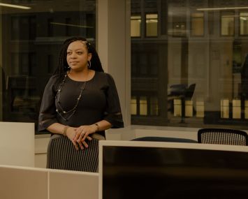 A woman with her hands folded standing among office desks.