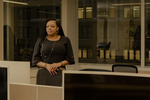 A woman with her hands folded standing among office desks.