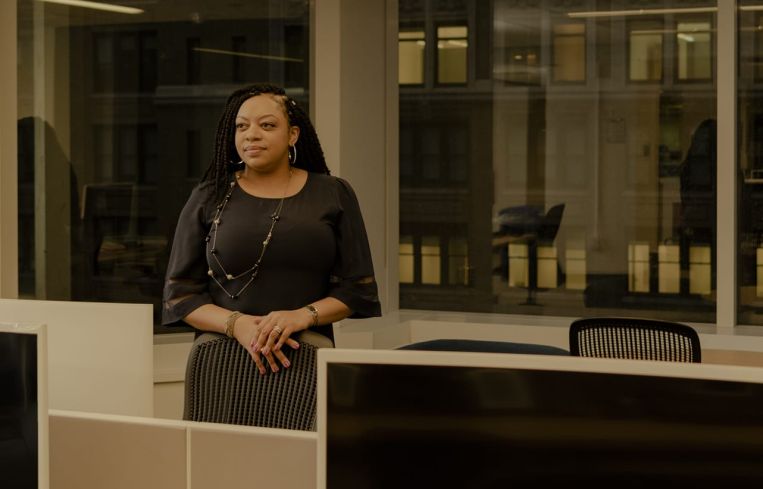 A woman with her hands folded standing among office desks.