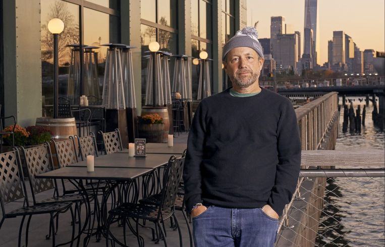 A man standing on a piece in front of some tables with tall buildings in the background.