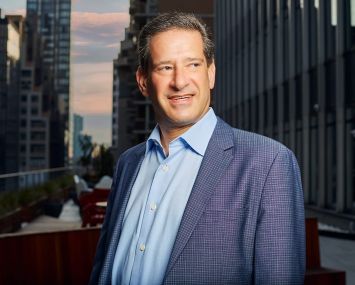 A man standing on a balcony with tall buildings behind him.