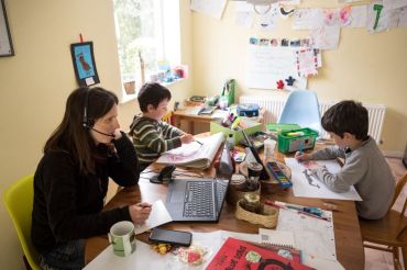 A mother works from surrounded by a children at a cluttered desk.