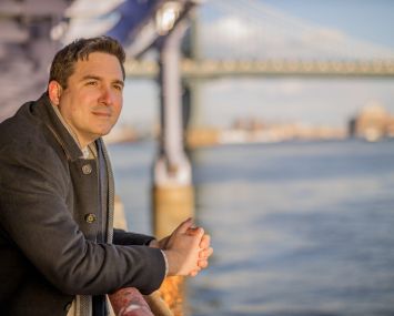 Ben Kallos overlooks the East River on a sunny day in New York. A bridge is visible behind him.