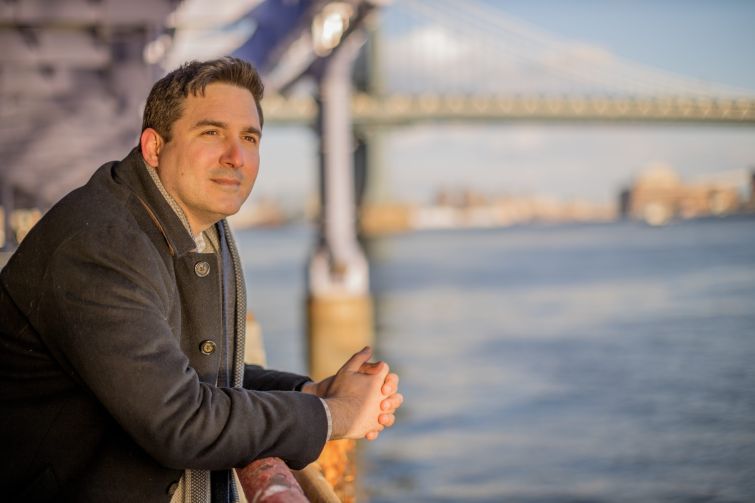 Ben Kallos overlooks the East River on a sunny day in New York. A bridge is visible behind him.