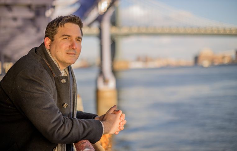 Ben Kallos overlooks the East River on a sunny day in New York. A bridge is visible behind him.