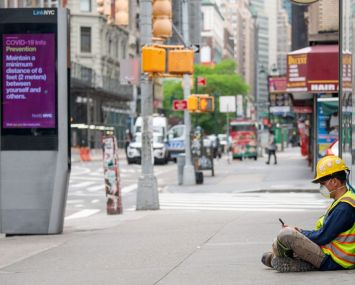 A construction worker wearing a mask sits on the street near a "COVID-19 info" digital display on May 28, 2020 in New York City.