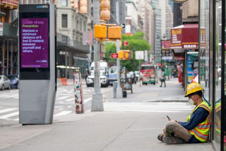 A construction worker wearing a mask sits on the street near a "COVID-19 info" digital display on May 28, 2020 in New York City.