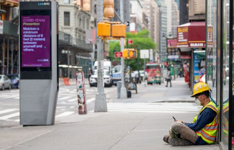 A construction worker wearing a mask sits on the street near a "COVID-19 info" digital display on May 28, 2020 in New York City.