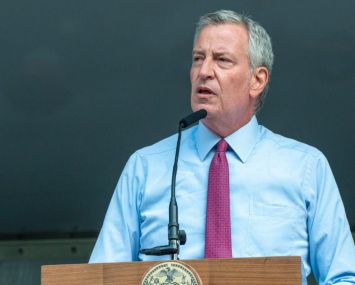 New York City Mayor Bill DeBlasio stands at a podium with a grey background behind him.