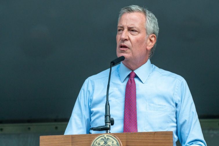 New York City Mayor Bill DeBlasio stands at a podium with a grey background behind him.