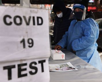 A sign saying "COVID 19 TEST" in front of a healthcare worker adorned in a face shield and blue suit.