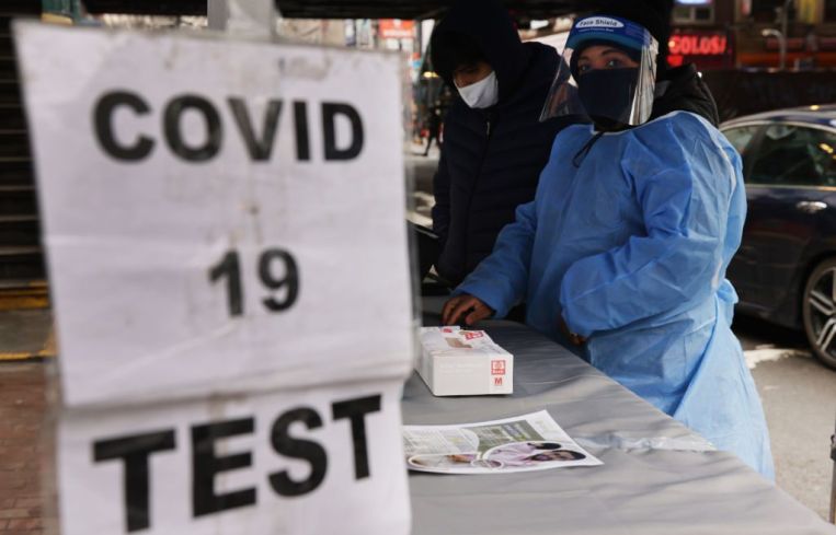 A sign saying "COVID 19 TEST" in front of a healthcare worker adorned in a face shield and blue suit.