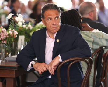 Governor of New York Andrew Cuomo sits at a decorated table at the Tribeca Festival looking off to the right, while figures in the background have their backs turned to him.