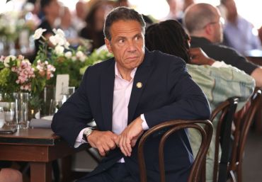 Governor of New York Andrew Cuomo sits at a decorated table at the Tribeca Festival looking off to the right, while figures in the background have their backs turned to him.
