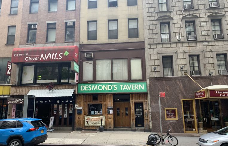 A grey storefront of an Irish Bar with a large green sign, named Desmond's Tavern.
