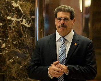 Matthew Calamari walks down a hallway with marble walls in sunglasses and a suit.