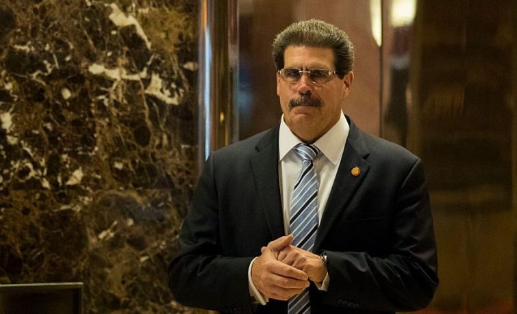 Matthew Calamari walks down a hallway with marble walls in sunglasses and a suit.