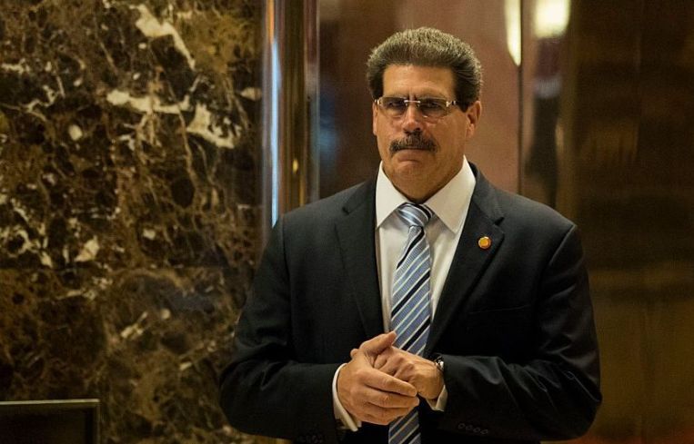 Matthew Calamari walks down a hallway with marble walls in sunglasses and a suit.