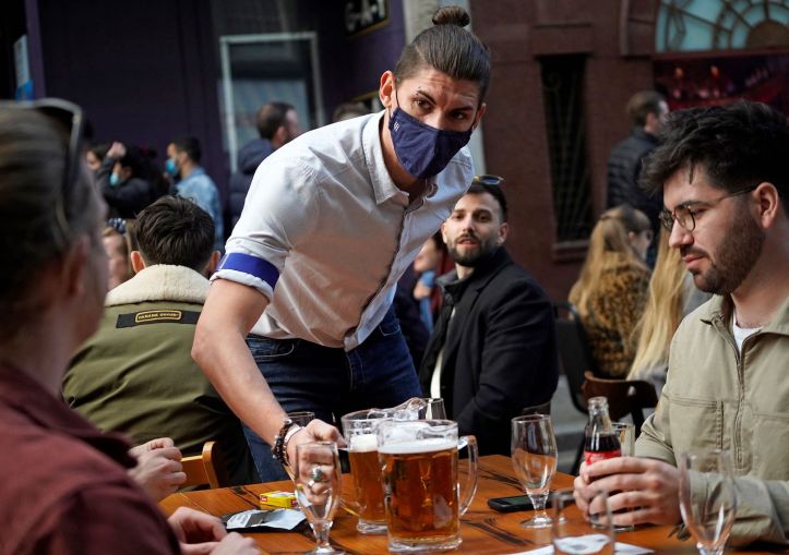 A hospitality worker serves a pitcher of beer to customers sat at an outdoor table of a re-opened bar.