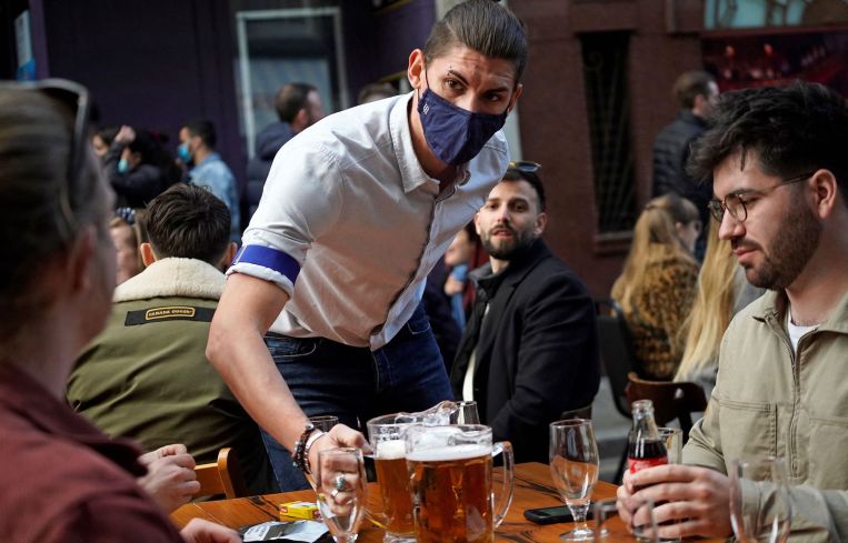 A hospitality worker serves a pitcher of beer to customers sat at an outdoor table of a re-opened bar.