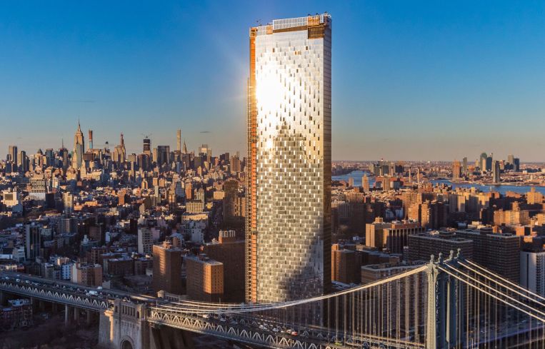 A large silver condo building rises above the Manhattan Bridge backed by a clear blue sky.