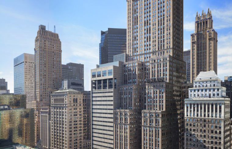 A tall, grey and brown skyscraper on a busy New York City block is backed by blue sky.