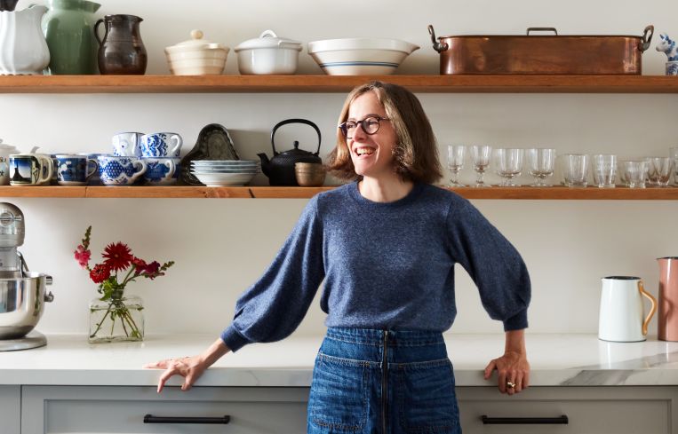 A woman smiling in front of a counter and shelves full of pots and pans.