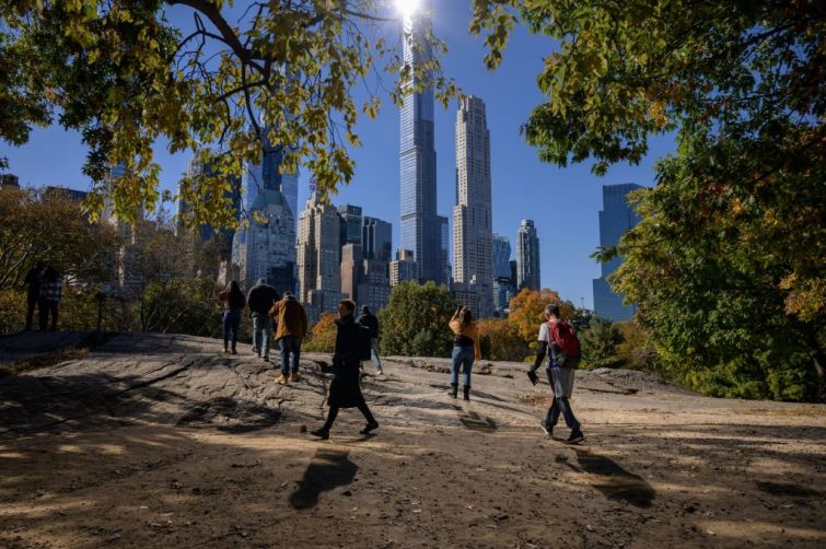 TOPSHOT - People stand beneath fall foliage and before high-rise buildings of the Manhattan city skyline in Central Park in New York on November 5, 2021. - merica's northeast is famous for its red, orange and yellow fall foliage but experts say climate change is dulling the colors and delaying peak season, causing concern for the region's multibillion-dollar "leaf-peeping" tourist industry. Warmer temperatures and heavier rainfall are keeping leaves greener for longer while extreme weather events like heatwaves and storms are stripping trees bare before getting to autumn, according to conservationists. (Photo by Ed JONES / AFP) (Photo by ED JONES/AFP via Getty Images)