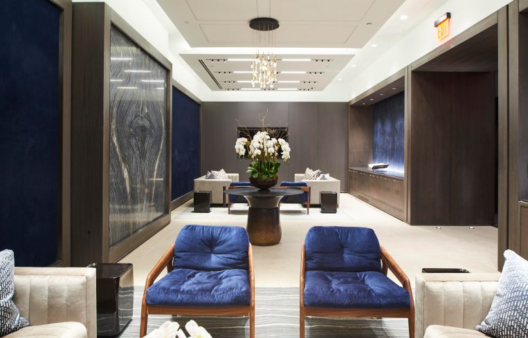 Two blue chairs lie in the forefront of a wide office room with a glass of flowers in the background.