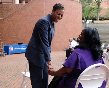 Queens Councilmember Adrienne Adams speaks with a seated woman outside a brick building.