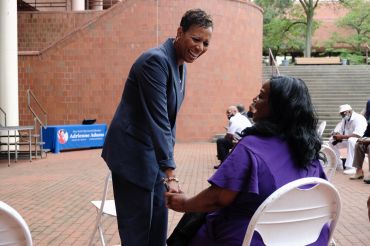 Queens Councilmember Adrienne Adams speaks with a seated woman outside a brick building.