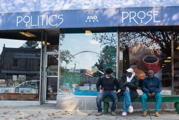Three men sit on a bench in front of the glass storefront of the Politics and Prose bookshop in Washington, DC on Nov. 30, 2017.