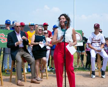 Rachel Loeb stands at a microphone ahead of a group of seated people at a baseball field
