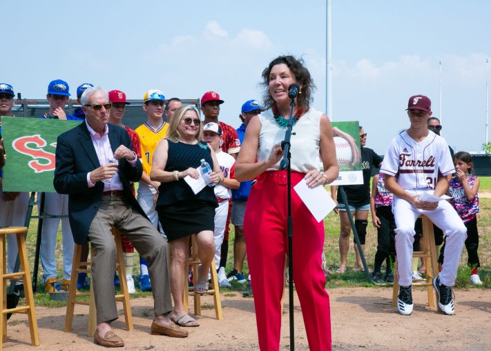 Rachel Loeb stands at a microphone ahead of a group of seated people at a baseball field