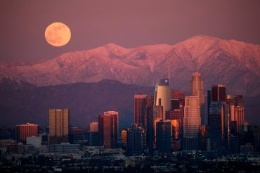 A full moon rises behind the snow-topped San Gabriel Mountains and the Downtown Los Angeles skyline at sunset as seen from the Kenneth Hahn State Recreation Area.