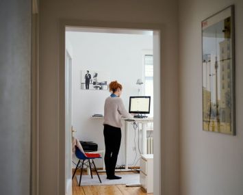 A woman is working in her apartment in front of a computer at a high desk.