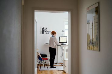 A woman is working in her apartment in front of a computer at a high desk.