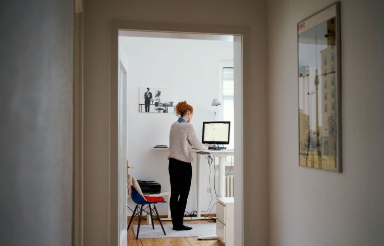 A woman is working in her apartment in front of a computer at a high desk.