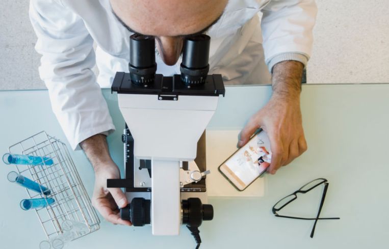 Mature scientist male in his 50s wearing a lab coat looking through a microscope in a laboratory. Basque Country, Spain, Europe.