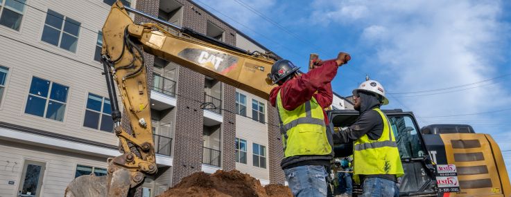Two workers at a construction site with a four-story building in the background.