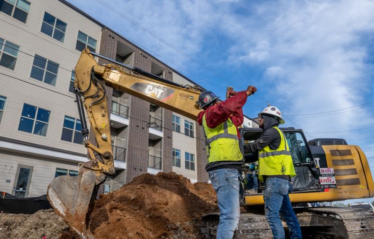 Two workers at a construction site with a four-story building in the background.