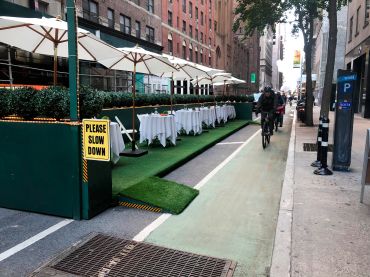 Outdoor Dining structure with Please Slow Down sign and bike lane filled with bike delivery people racing by, Manhattan, New York.