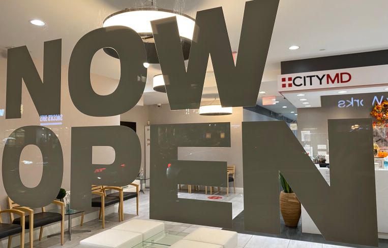 The front desk of a health clinic seen through a storefront window with the words "now open."