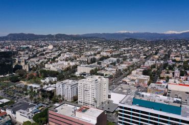 An aerial image shows snow capped mountains and the Hollywood sign on the horizon behind a view of Los Angeles neighborhoods including Westlake and East Hollywood.