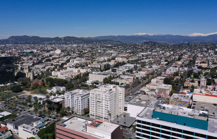 An aerial image shows snow capped mountains and the Hollywood sign on the horizon behind a view of Los Angeles neighborhoods including Westlake and East Hollywood.