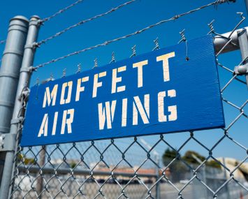 Blue and white stenciled sign for the Moffett Air Wing hanging on a barbed wire fence at Moffett Field, within the secure area of the NASA Ames Research Center campus in Mountain View, Calif., in 2016.