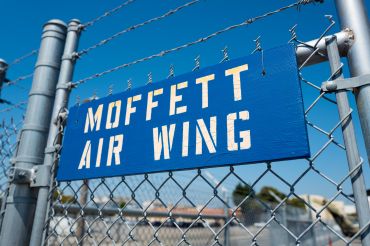 Blue and white stenciled sign for the Moffett Air Wing hanging on a barbed wire fence at Moffett Field, within the secure area of the NASA Ames Research Center campus in Mountain View, Calif., in 2016.