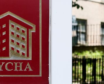 A NYCHA sign in front of a patch of grass with a person walking into a housing development in the background.